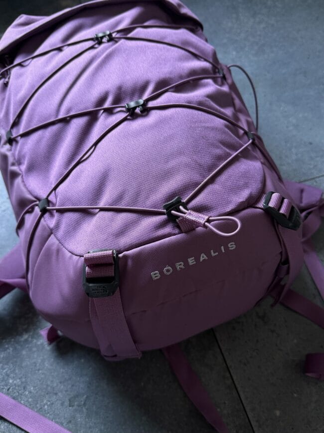 Purple Borealis backpack with exterior bungee cords and buckles resting on a dark floor.