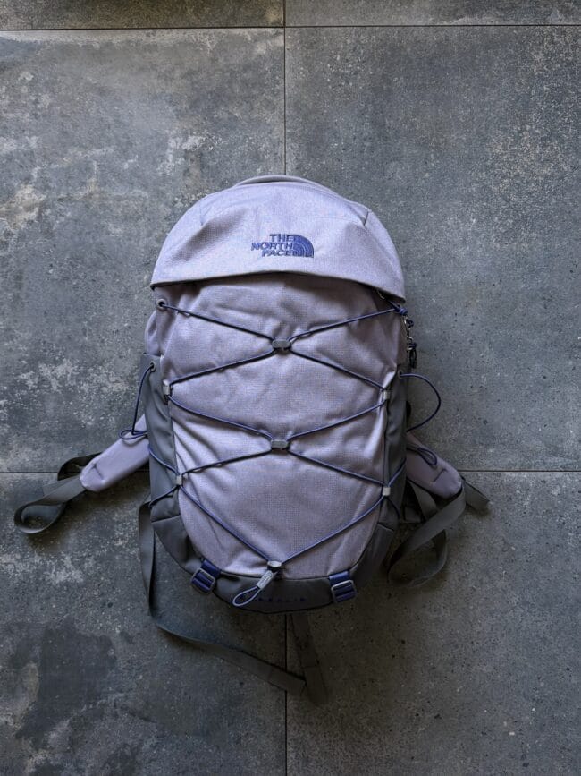 Grey North Face backpack with bungee cords lying on a concrete surface, front view showing logo and straps.