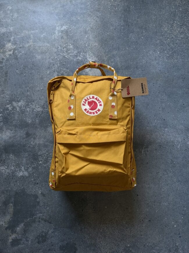Mustard-yellow Fjällräven backpack with polka-dot straps and a product tag on a blue concrete background.