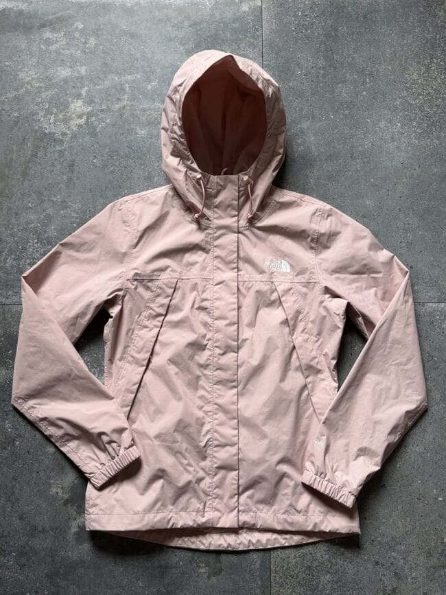 Beige hooded rain jacket laid flat on a concrete floor, North Face logo on the chest area.