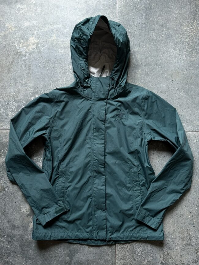 Teal green hooded jacket laid flat on a gray concrete floor, showing The North Face logo on the chest area.