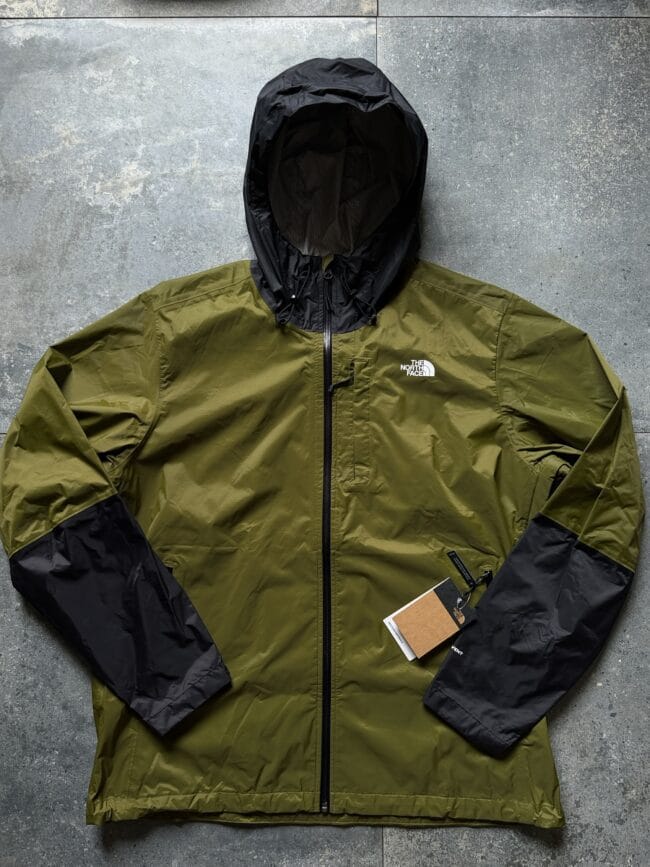 Olive green The North Face rain jacket laid flat with a black hood and sleeves, front zipper visible on a gray concrete background