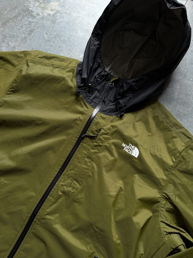 Olive-green The North Face jacket with a black hood and front zipper, laid flat.
