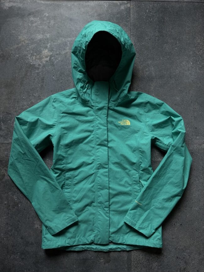 Green North Face jacket laid flat on a dark stone floor, hood up, full front view.