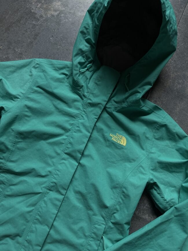 Teal-green The North Face jacket with hood up, laid flat and logo visible on the chest.