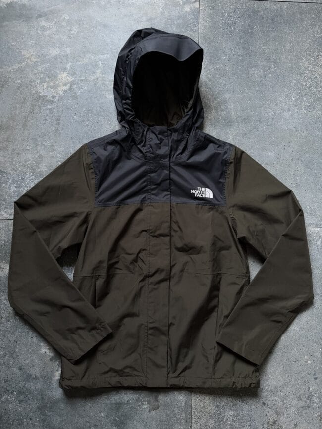 Two-tone hooded jacket (black and olive) laid flat on a gray concrete surface, The North Face logo visible on the chest pocket area on the right side of the image.