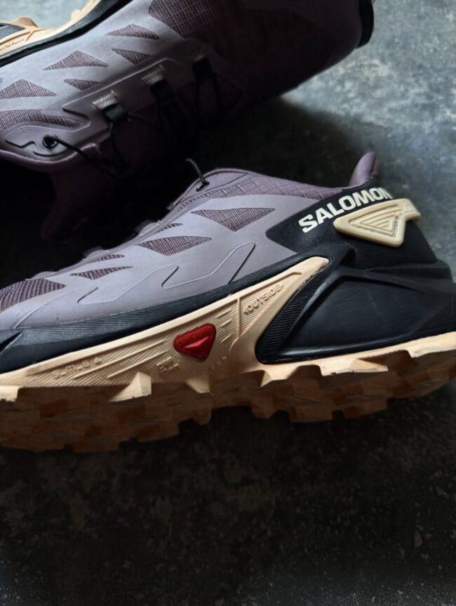 Close-up of a Salomon trail running shoe showing a gray upper, black heel, and beige rugged outsole with chunky tread.