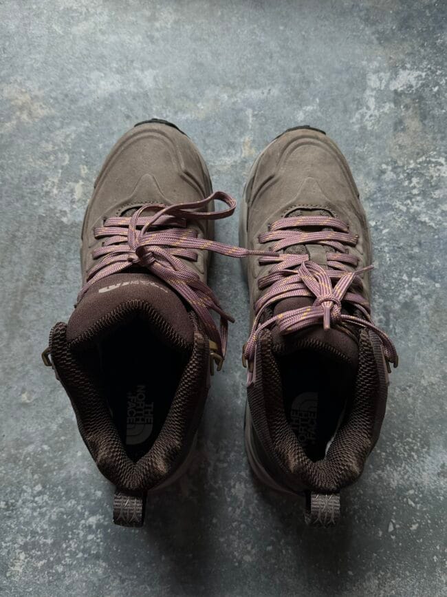 Top-down view of a pair of brown hiking boots with purple laces on a gray concrete floor, toes pointing outward.