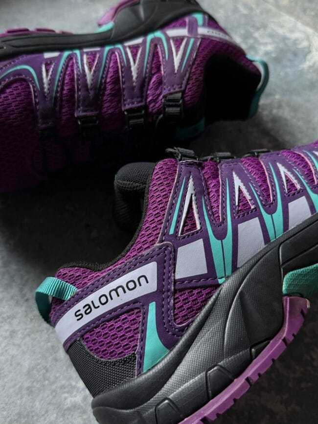 Close-up of a purple Salomon trail shoe with teal accents and a black sole on a gray background.