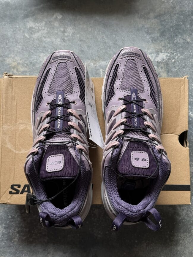 Top-down view of two purple Salomon trekking shoes resting on a cardboard shoebox, laces tied, showing mesh and synthetic overlays.
