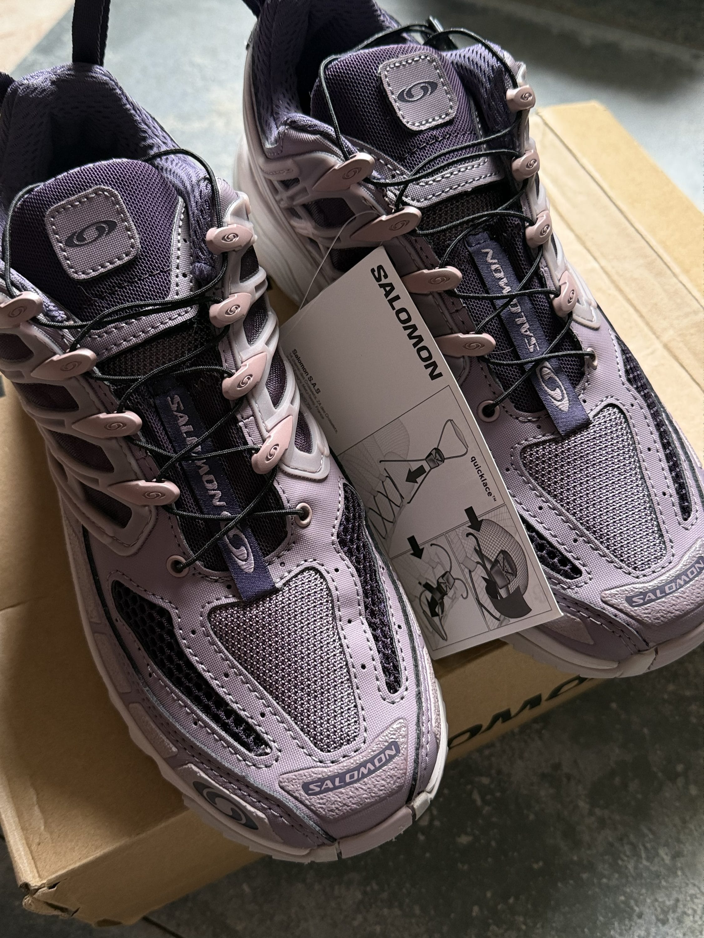 Pair of lavender Salomon hiking shoes with tan laces, placed on a cardboard box with a product tag attached.