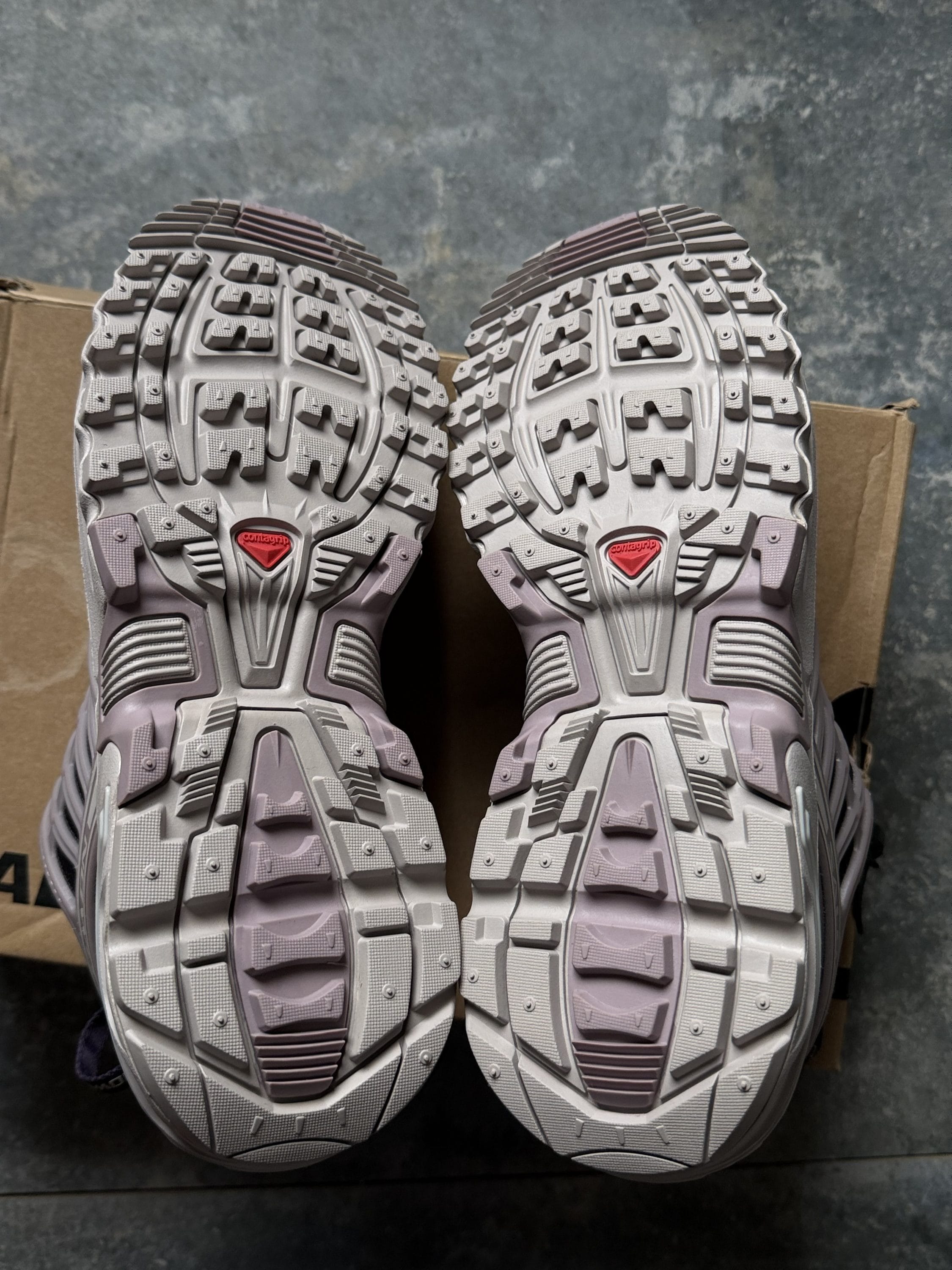 Pair of grey and lavender hiking boots viewed from above, highlighting their rugged tread soles on a boxy surface