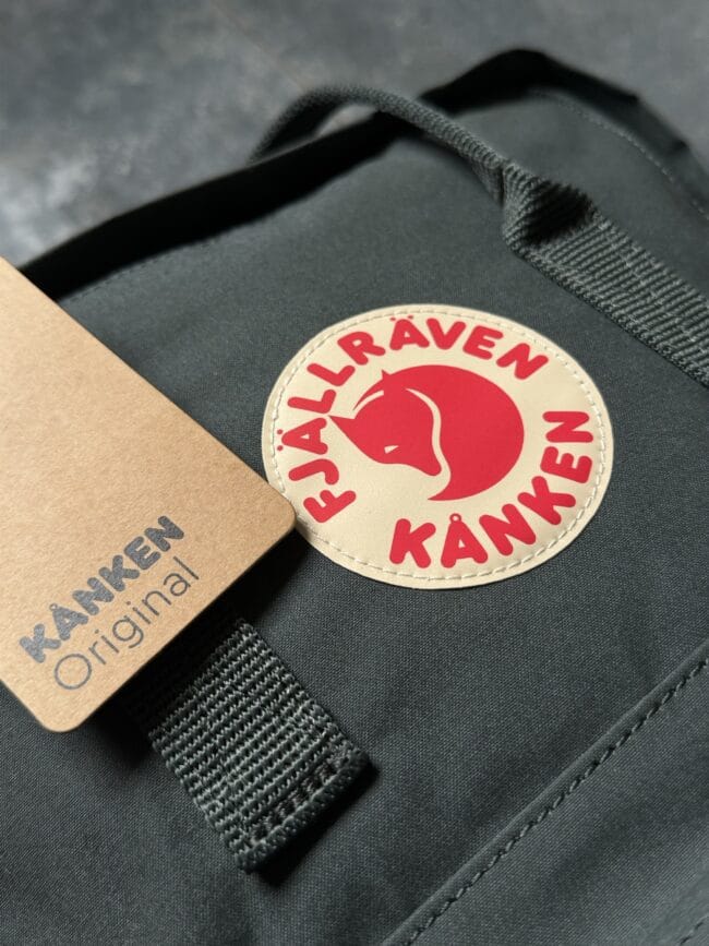 Close-up of a dark Fjällräven Kånken backpack showing a beige circular logo patch with a red fox and KÅNKEN text, with a brown tag nearby and black straps/rivets.