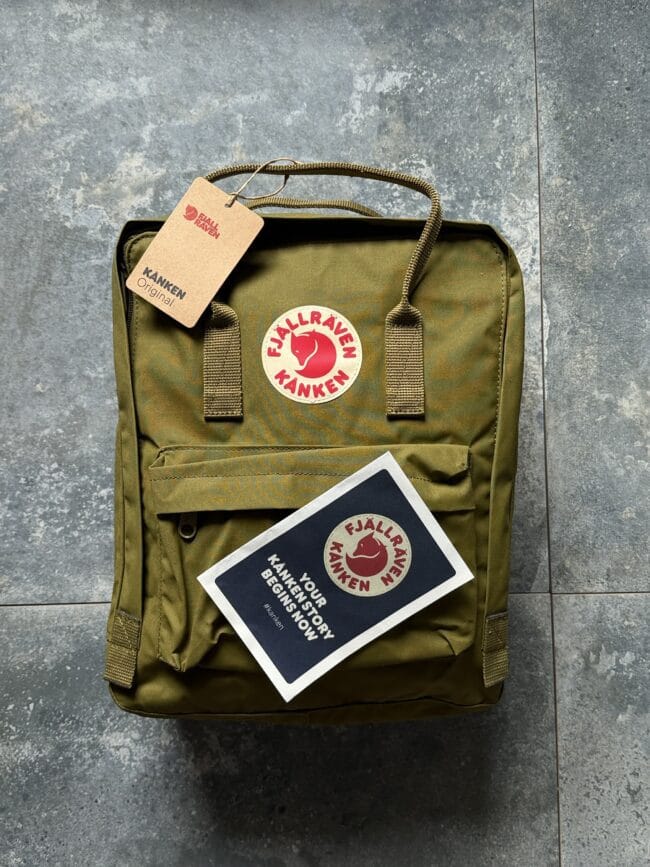 Olive Kanken backpack with round red Fjällräven logo patch and attached brown tag on a gray tiled surface.