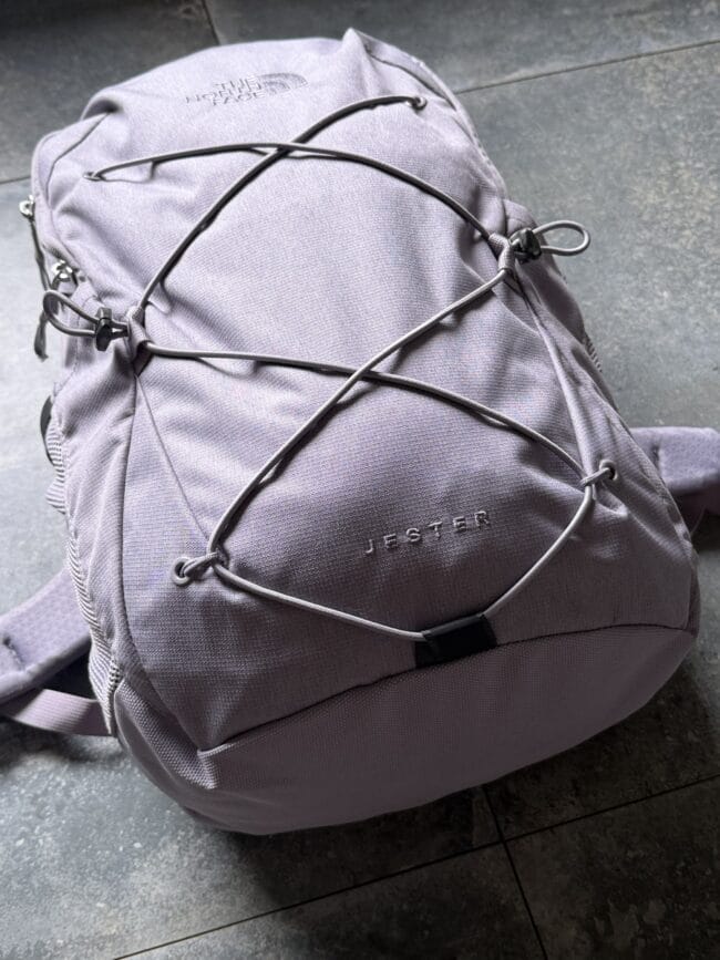 Gray The North Face Jester backpack with cross‑hatched bungee cords on a dark tiled floor.
