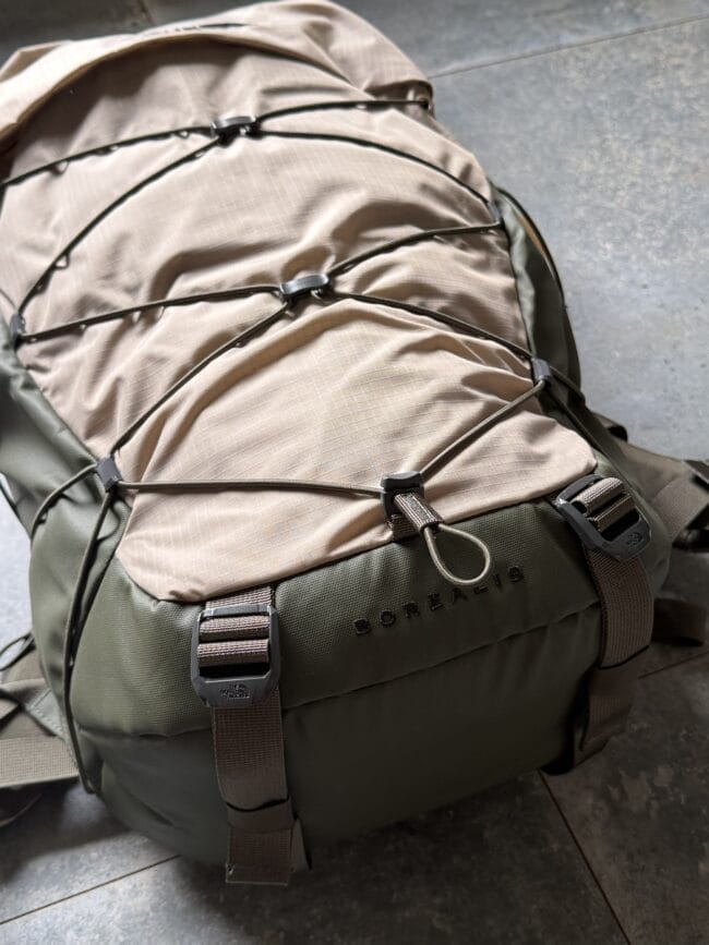 Olive backpack close-up with bungee cords and straps, showing brand hardware and rugged outdoor design.