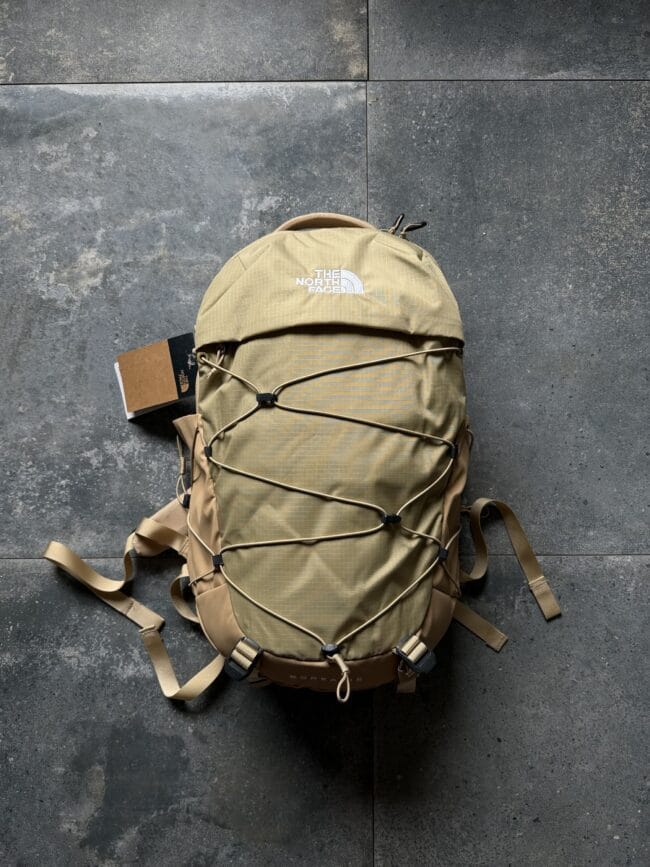Tan The North Face backpack lying on a dark gray tiled floor, with external bungee cords and straps visible.
