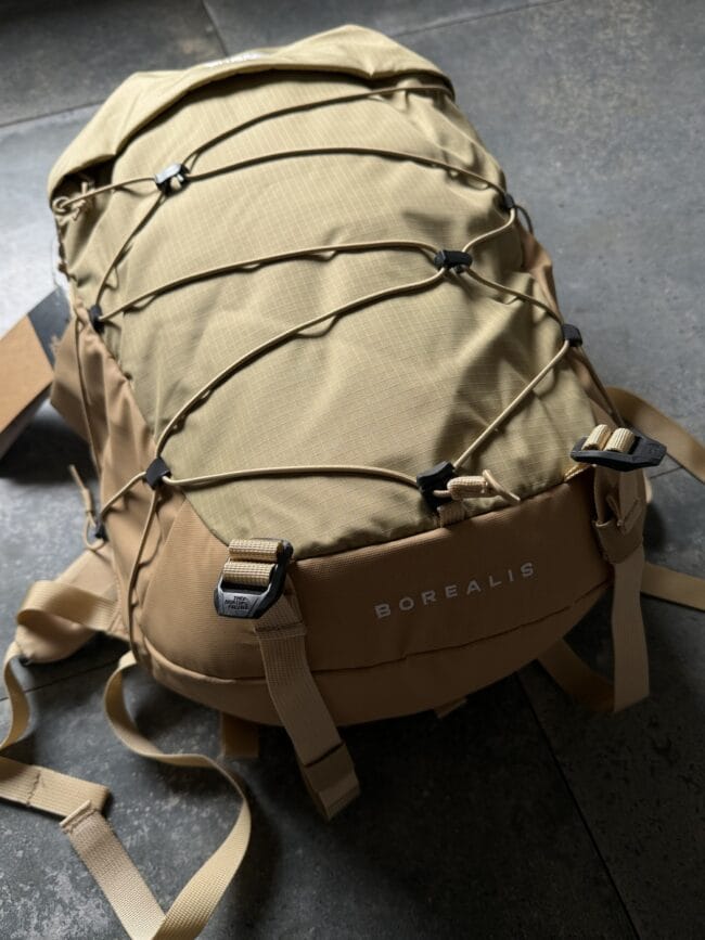 Beige Borealis tactical backpack with external bungee cords and multiple straps laid on a dark floor.