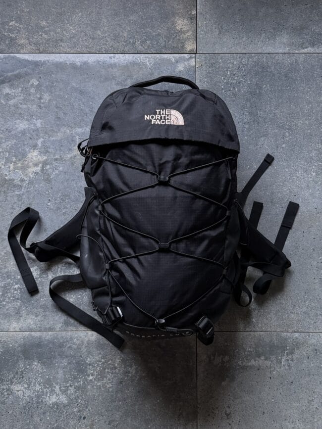 Black The North Face hiking backpack lying on a gray tiled floor with bungee straps and side straps visible.