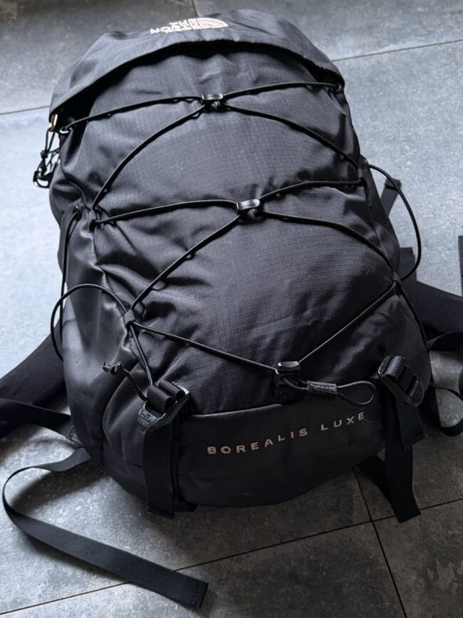 Black Borealis Luxe backpack with crisscross bungee cords on a gray tiled floor.