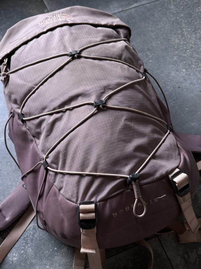 Brown hiking backpack with external beige bungee cords and plastic cord locks, resting on a dark tiled surface. The North Face logo is visible on the top flap.