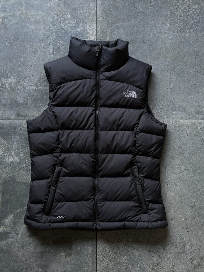 Black puffer vest by The North Face with front zipper, displayed against a gray stone background.