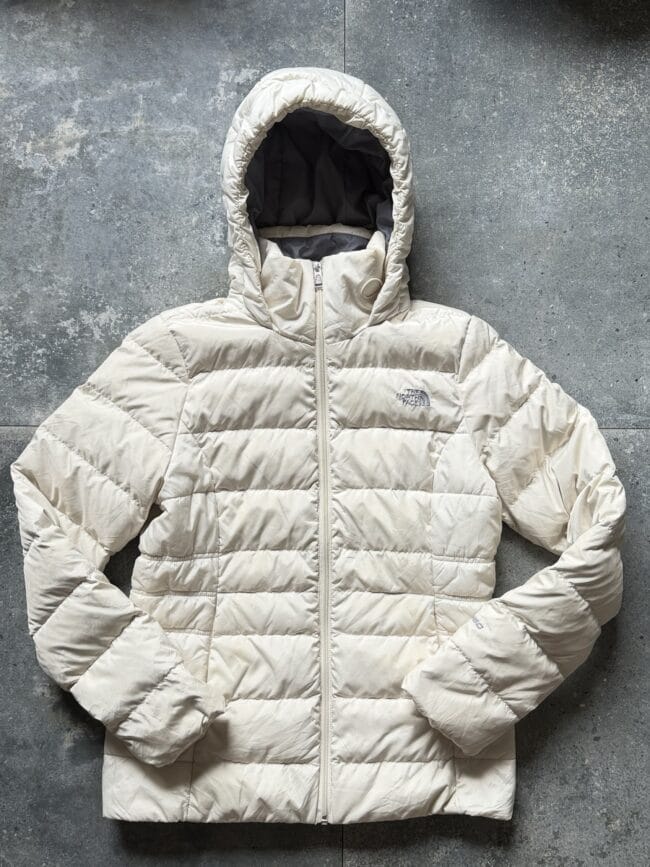 Beige puffer jacket with hood laid flat on a gray concrete surface, zipper closed, logo on chest (The North Face).