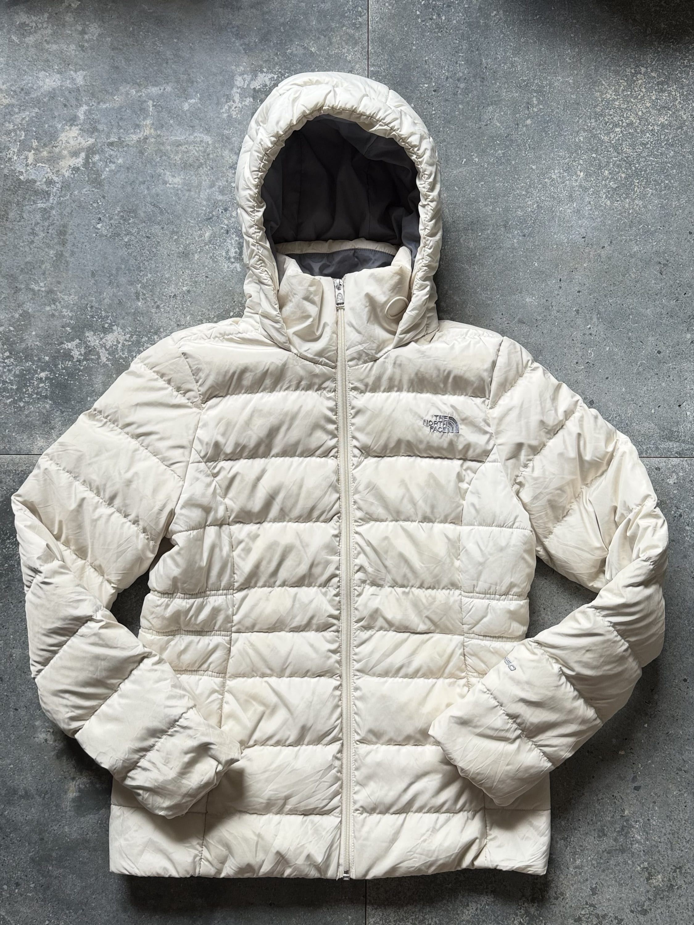 Beige puffer jacket with hood laid flat on a gray concrete surface, zipper closed, logo on chest (The North Face).