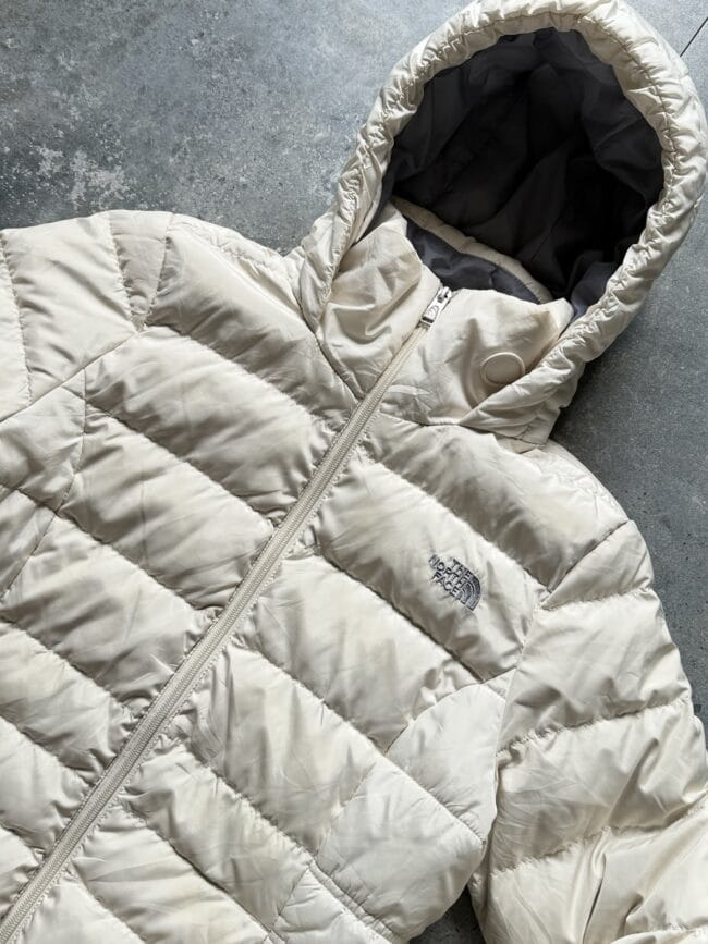 Beige quilted puffer jacket with hood, full-front zipper, lying on a gray surface; The North Face logo visible on the chest area.