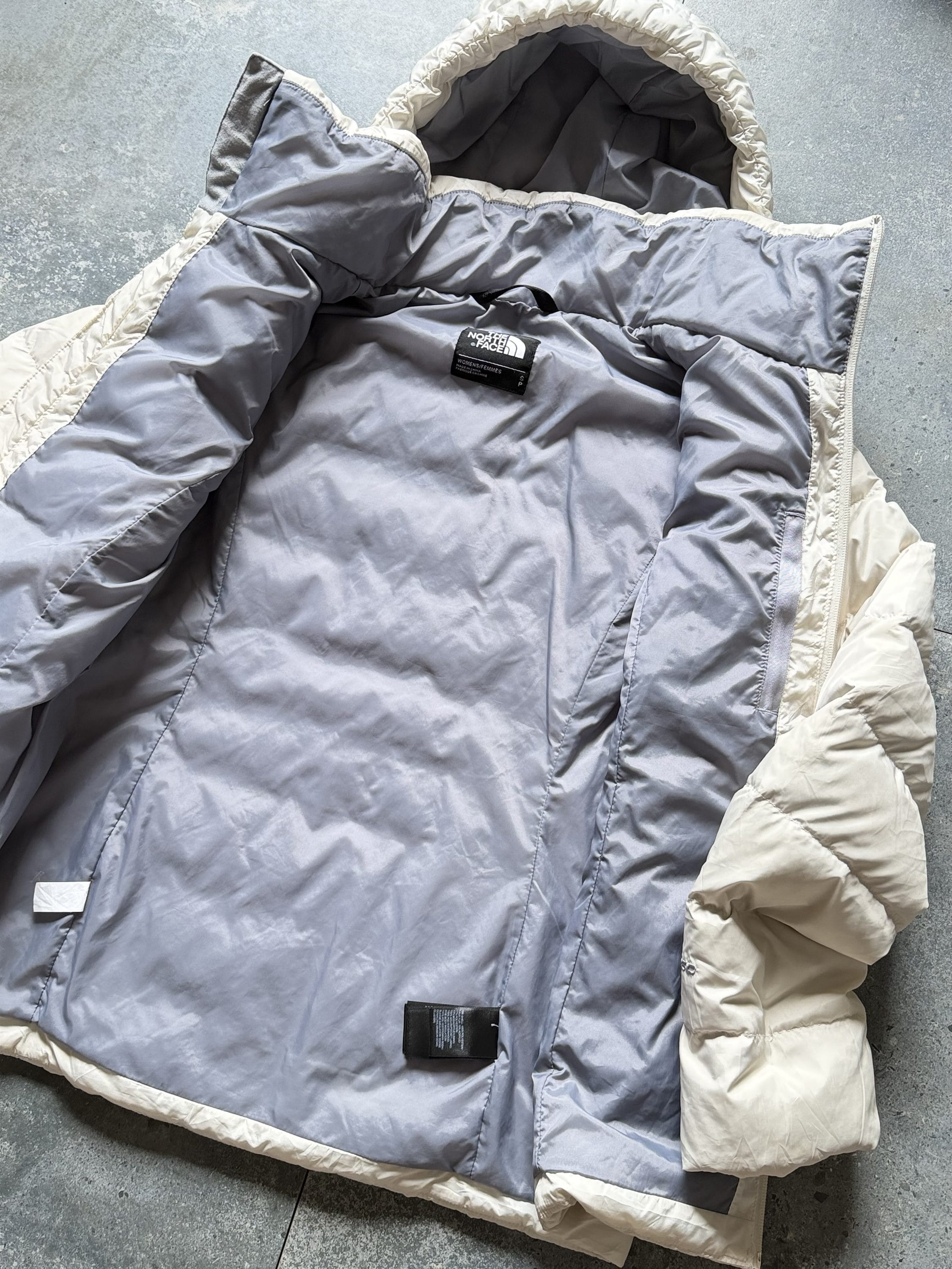 Open North Face puffer jacket showing its light gray lining and beige exterior on a concrete floor, interior visible beyond the zipper edge.