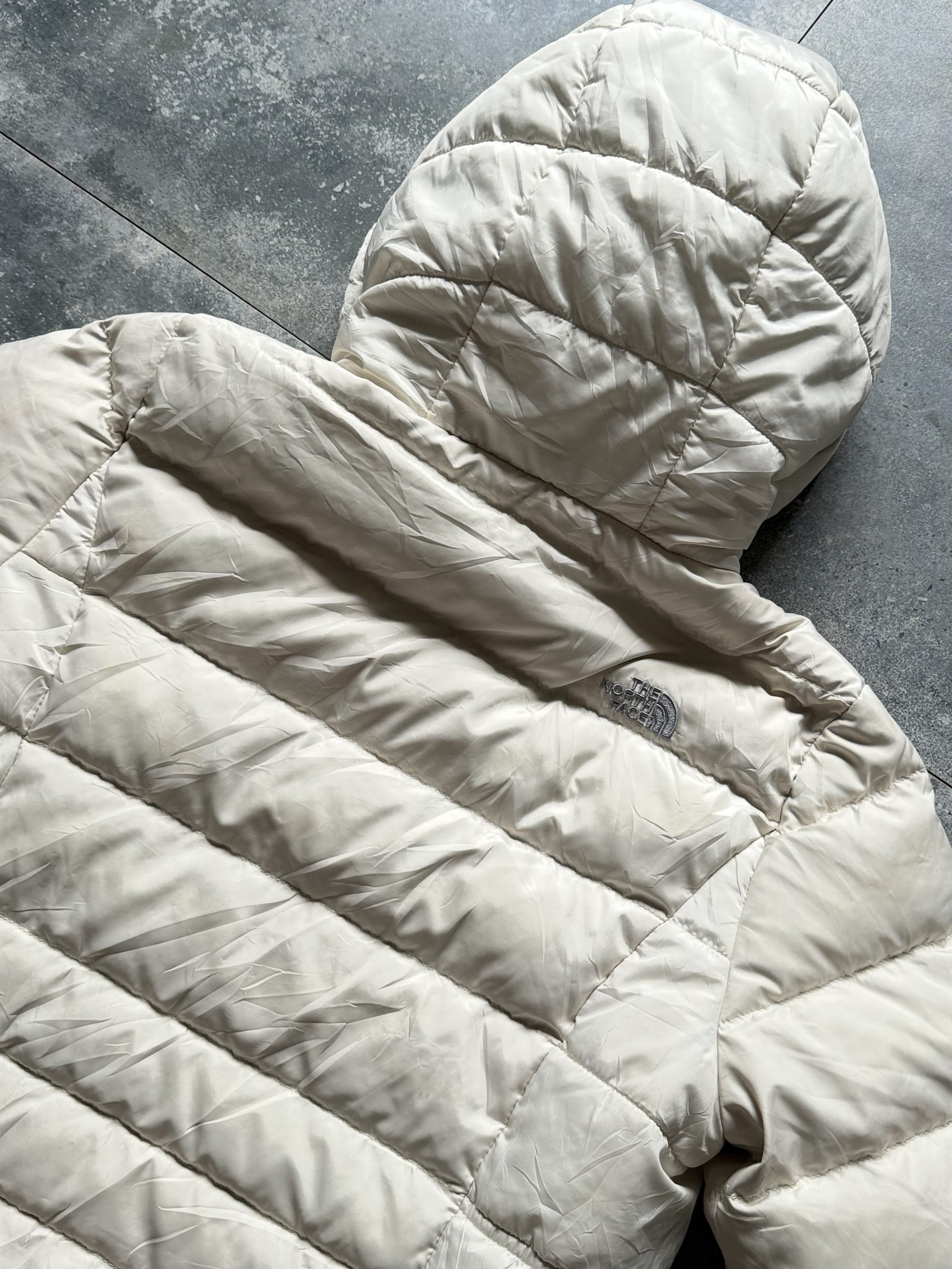 Beige quilted puffer jacket laid on a gray concrete floor, with the North Face logo visible on the side pocket area.