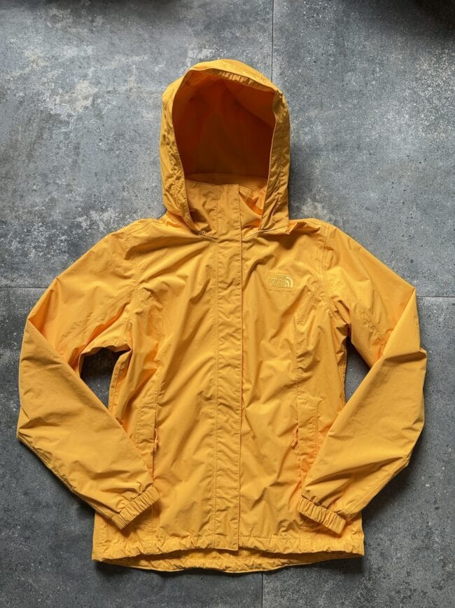 Orange hooded waterproof jacket laid flat on a gray concrete surface; front zipper and elastic cuffs visible, brand logo on chest (The North Face).