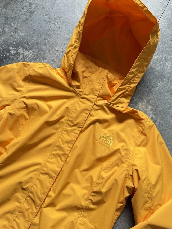 Orange The North Face rain jacket laid flat on a gray concrete surface, hood up, showing outer material and stitching details.