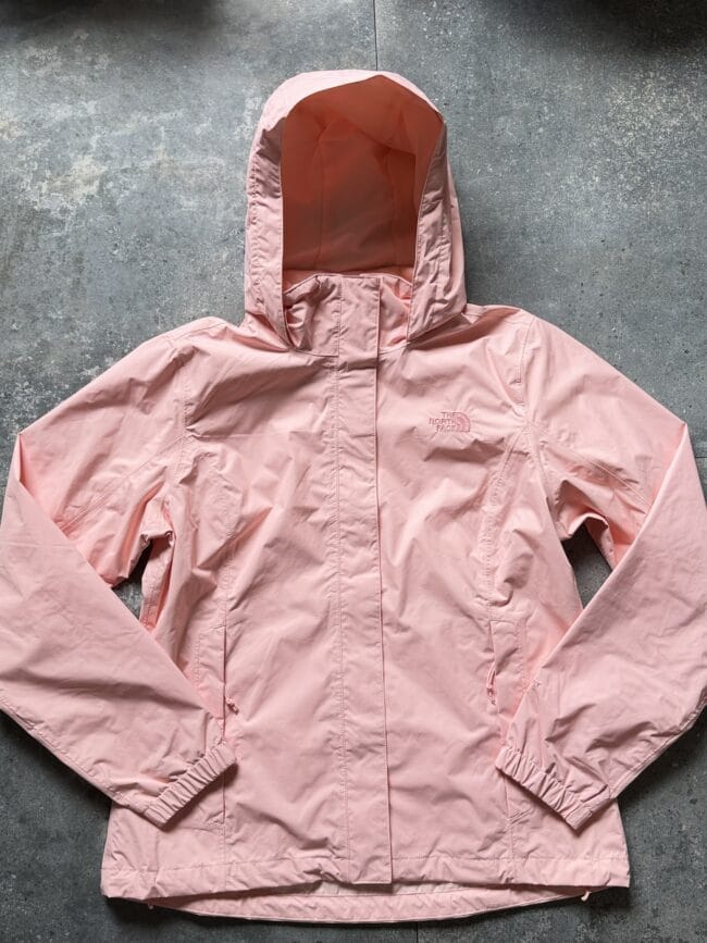 Light pink hooded jacket laid flat on a gray concrete surface, with the North Face logo on the chest.