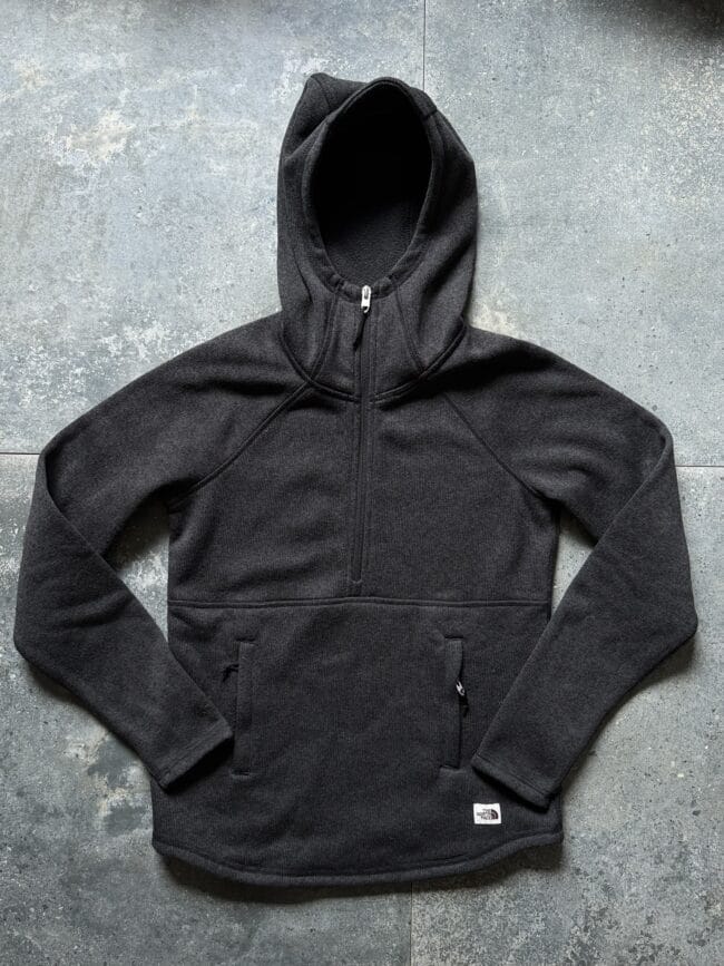 Black hoodie laid flat on a gray concrete floor, zipper closed and a small North Face tag near the hem.