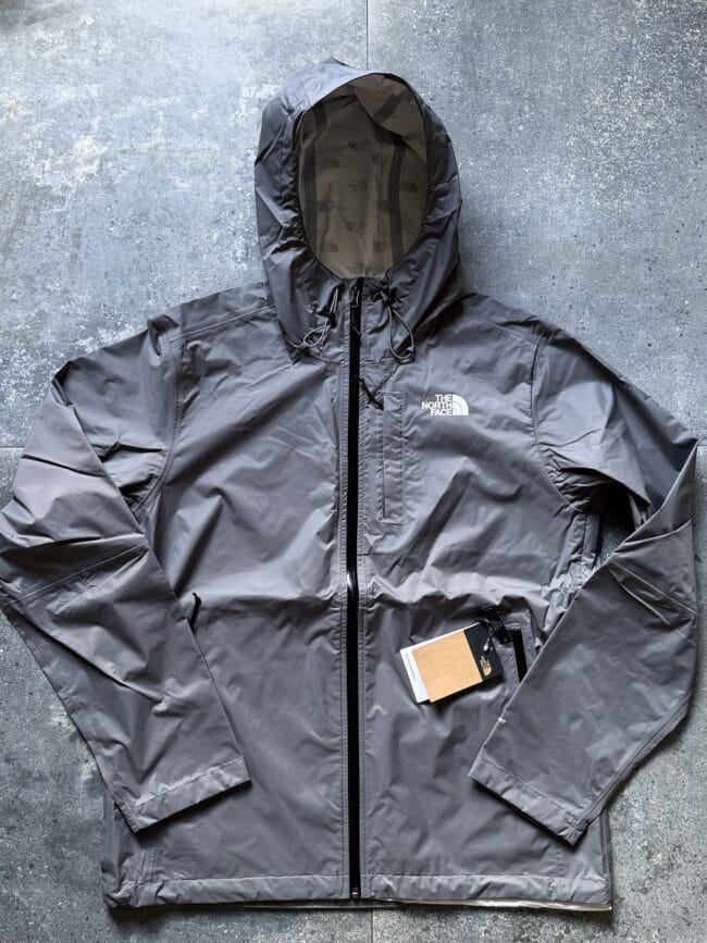 Gray The North Face hooded jacket laid flat on a concrete surface; front zipper, drawstring hood, logo on the chest, and attached tags.