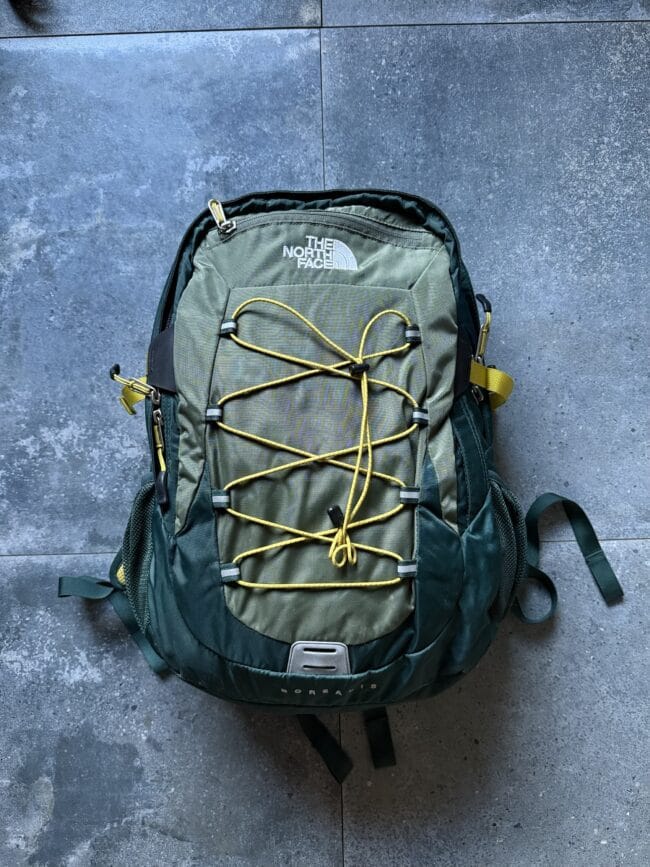 Green North Face hiking backpack with yellow bungee cords on the front, resting on a gray tiled surface.