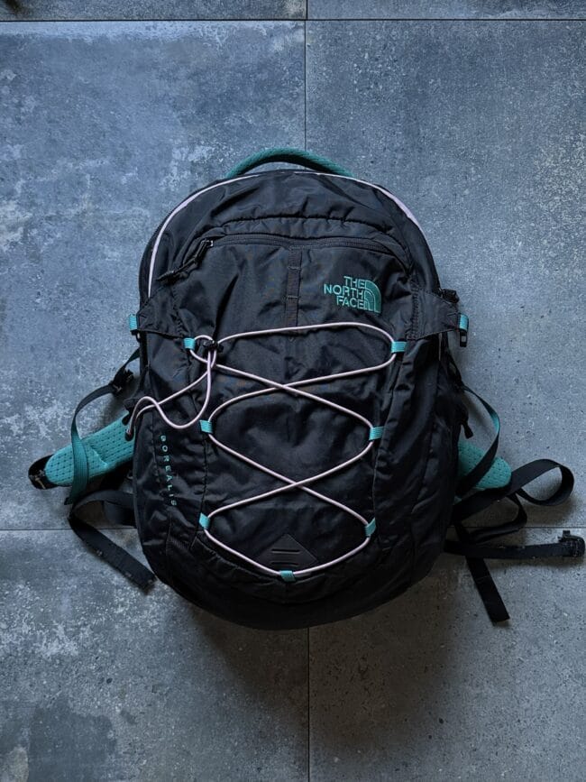 Dark backpack with light teal straps and bungee cords, laid on a gray stone floor; shows The North Face logo on the front pocket