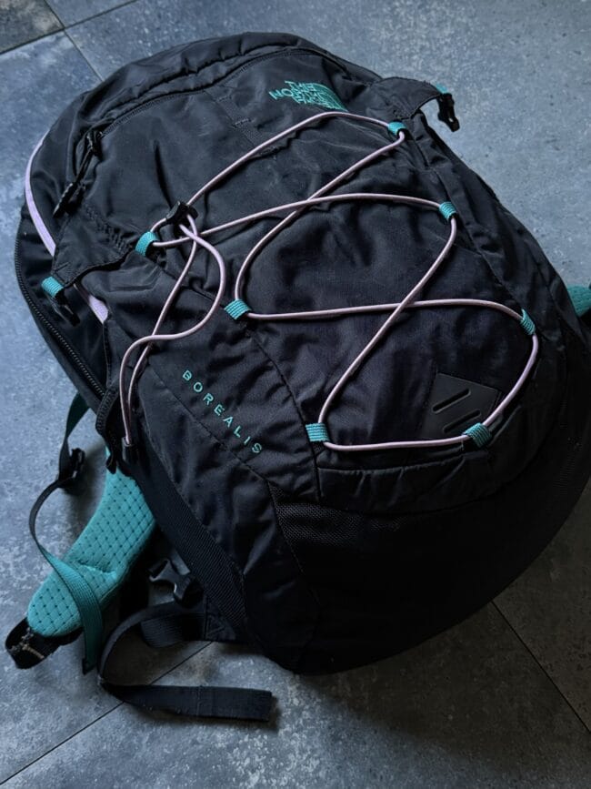 Black North Face backpack on a gray tiled floor, with pink bungee cords and teal straps visible on the outside.