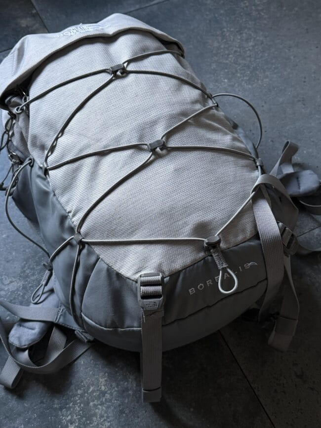 Grey The North Face backpack with exterior bungee cords and straps on a dark floor surface.