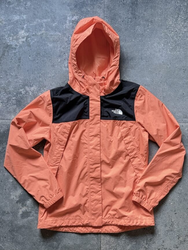 Orange-pink The North Face windbreaker with black chest panel and hood laid on a gray concrete floor.
