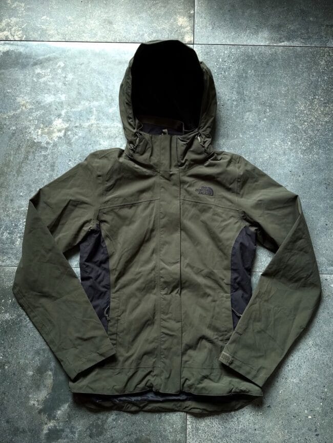 Olive green North Face hooded waterproof jacket laid flat on a textured concrete surface, showing front zipper and pockets.