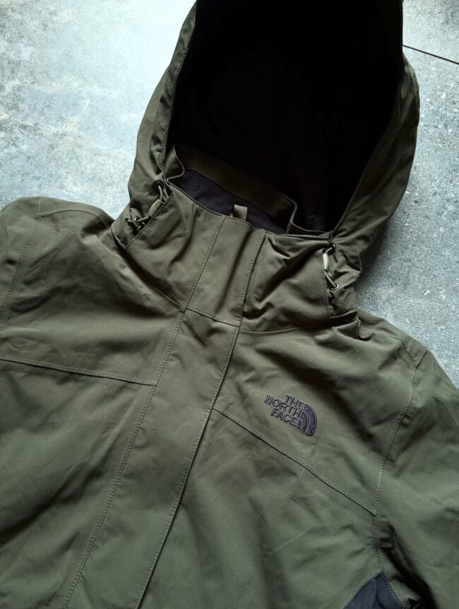 Olive green North Face rain jacket with hood open, showing black inner lining and visible stitching on a gray surface.