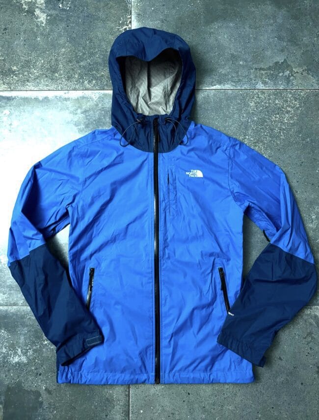 Blue North Face rain jacket with hood up, full front zipper closed, lying on a gray concrete floor with a textured surface.