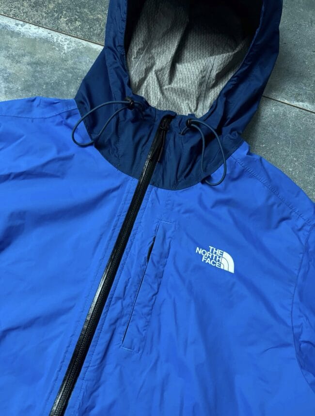 Blue North Face jacket with hood and front zipper, close-up detail of drawstrings and logo visible on chest