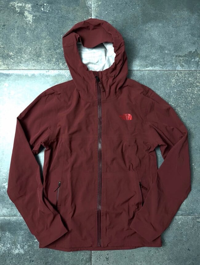 Maroon North Face hooded windbreaker laid flat on a textured tile floor with front zipper and logo on the chest.