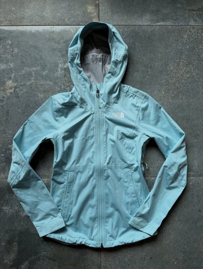 Light blue waterproof jacket with hood laid flat on a dark gray stone surface, zipper closed and North Face logo visible on the chest.