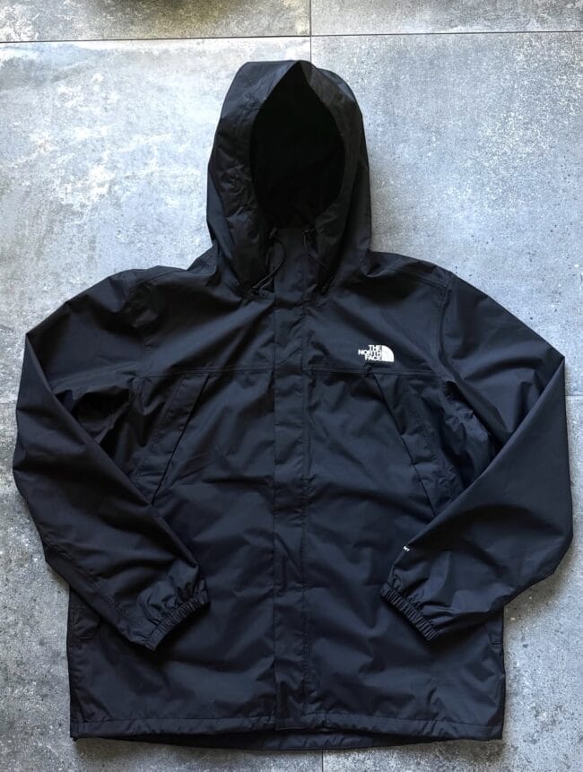 Black waterproof hooded jacket laid flat on a concrete floor, with The North Face logo visible on the chest area.