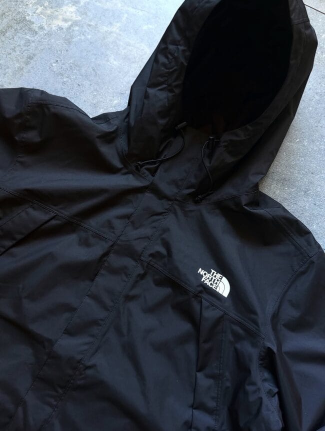 Black North Face hooded rain jacket laid flat on a gray concrete surface, showing the logo on the chest pocket area.