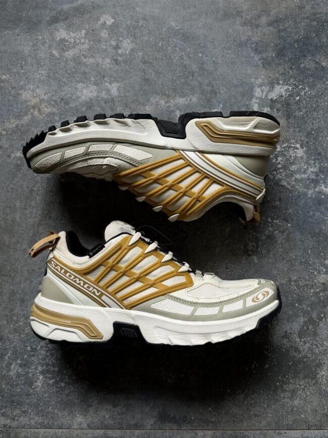 Pair of white and beige Salomon trail running shoes with gold accents photographed from side on a textured gray surface, showing rugged outsole.